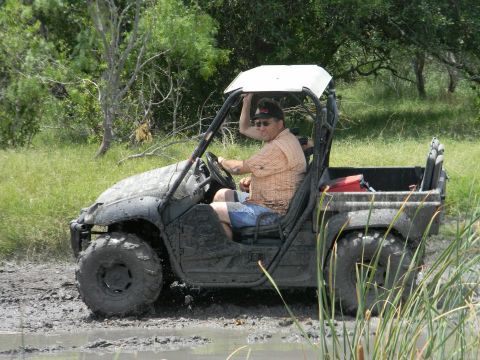 1989 Ford Bronco - A man drives a side-by-side UTV through a muddy, off-road area with tall grass.