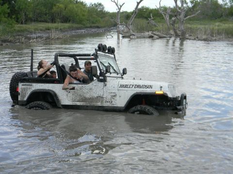 1989 Ford Bronco - A white Jeep Wrangler with 'Harley Davidson' branding navigates through muddy water with several occupants.