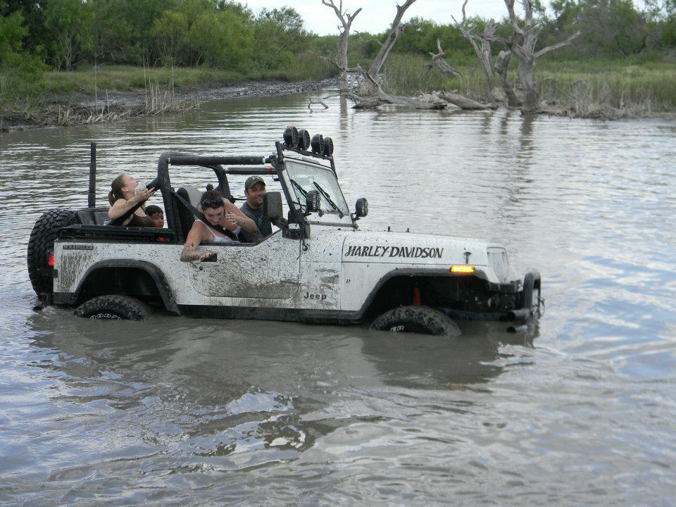 White Jeep Wrangler with 'Harley Davidson' decal stuck in muddy water, people inside