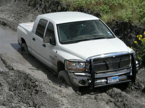 1989 Ford Bronco - A white pickup truck is stuck in thick mud, with its tires submerged and the front end angled downwards.