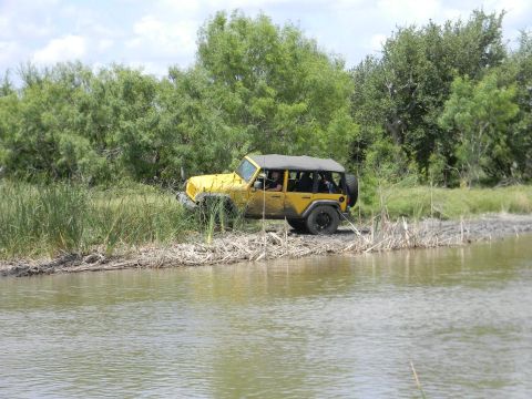 1989 Ford Bronco - Yellow Jeep Wrangler parked on a muddy bank by the water, with reeds and trees in the background.