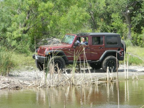 1989 Ford Bronco - A red Jeep Wrangler is parked near the edge of a body of water, surrounded by vegetation.