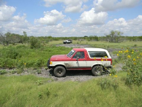1989 Ford Bronco - A red and white Ford Bronco navigates a muddy off-road path under a cloudy sky, with another vehicle visible in the distance.