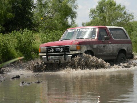 1989 Ford Bronco - This red and grey Ford Bronco navigates through a muddy water crossing, kicking up a large splash.