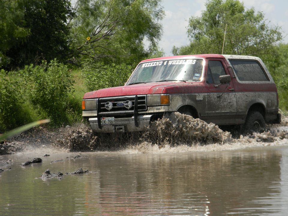 Red and grey Ford Bronco driving through mud, front grille visible, windshield sticker reads WEEKEND 4 WHEELERS