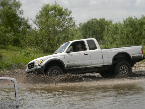 1989 Ford Bronco - A white Toyota Tacoma navigates a muddy water crossing with trees in the background.
