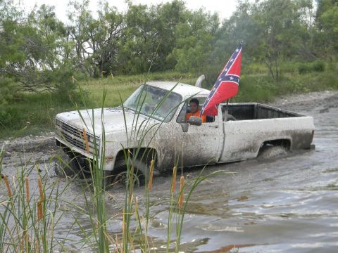 1989 Ford Bronco - A white pickup truck is stuck in the mud, with a person inside and a confederate flag flying from the bed. The grille indicates it is likel…