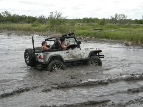 1989 Ford Bronco - Two people are riding in a white Jeep Wrangler as it drives through a muddy, watery area.