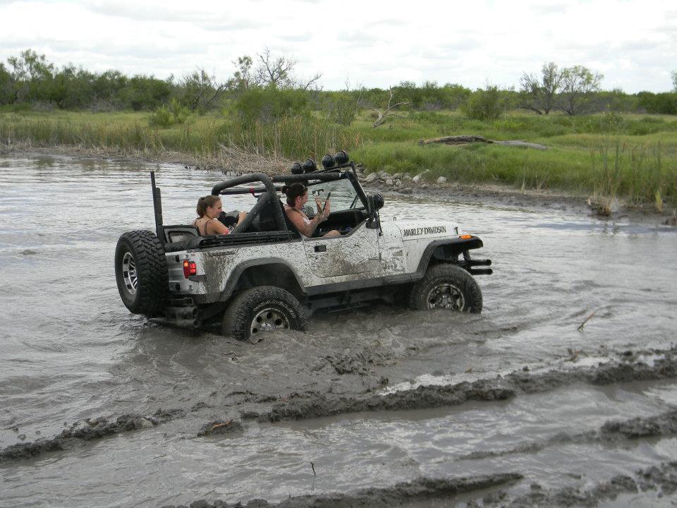White Jeep Wrangler with two people inside driving through a muddy water area. Harley-Davidson text visible on the side