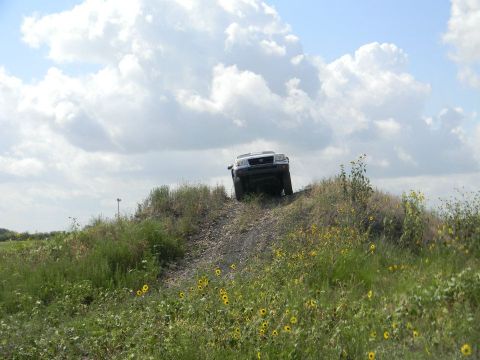 1989 Ford Bronco - A white SUV navigates an off-road trail, climbing a steep dirt incline under a cloudy sky.
