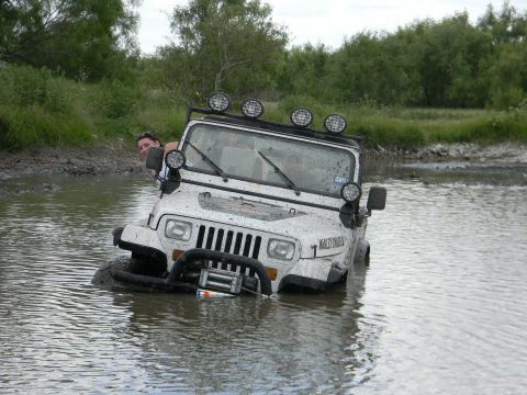 1989 Ford Bronco - A white Jeep Wrangler is partially submerged in muddy water, with a person visible inside.