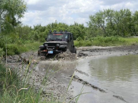 1989 Ford Bronco - A black Jeep Wrangler navigates a muddy trail, kicking up water and dirt.