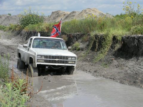 1989 Ford Bronco - White pickup truck navigates a muddy track, flying a Confederate flag.