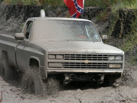 1989 Ford Bronco - A mud-splattered pickup truck navigates a muddy track, with a Confederate flag flying in the background.