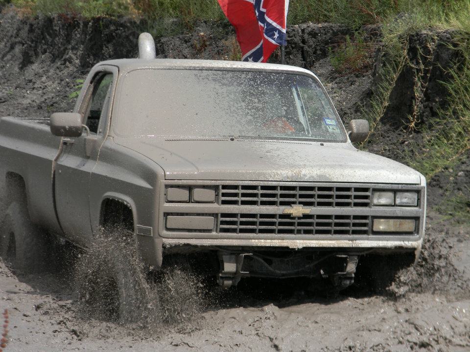 Mud-covered pickup truck driving through mud, front view. A Confederate flag is visible