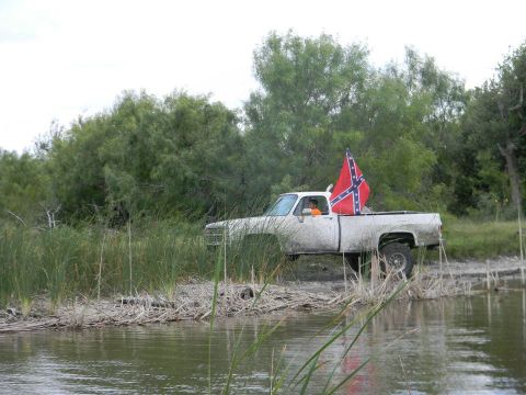 1989 Ford Bronco - A white pickup truck is partially submerged in water, with a Confederate flag flying from its bed.