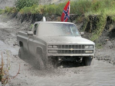 1989 Ford Bronco - A mud-splattered Chevrolet pickup truck navigates a muddy terrain, flying a Confederate flag.