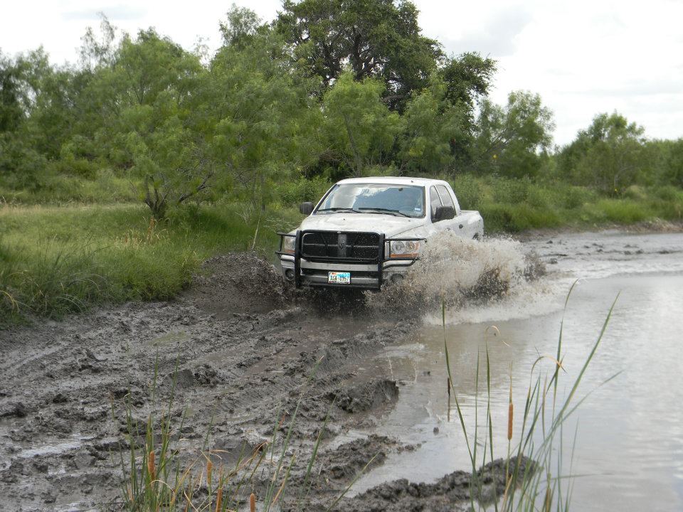 White pickup truck splashing through mud and water, trees and brush in background