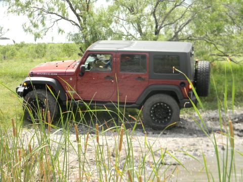1989 Ford Bronco - A red Jeep Wrangler navigates through a muddy, grassy terrain with trees in the background.