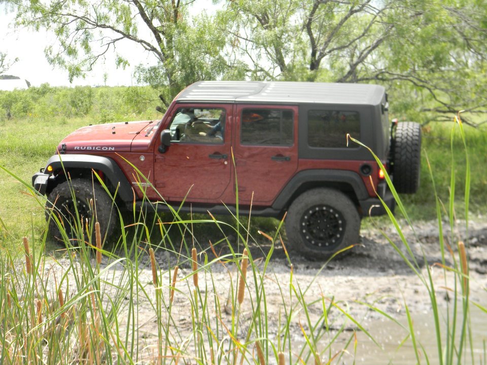 Red Jeep Wrangler with a soft top, driving through mud and tall grass