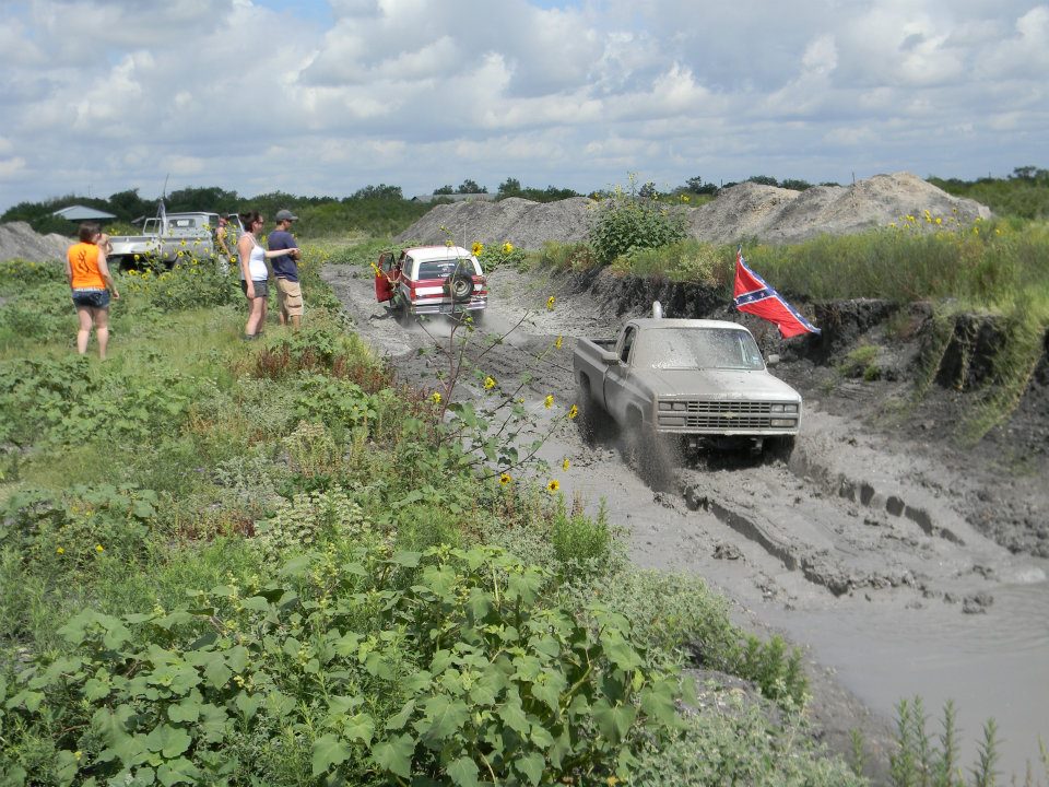 Silver pickup truck driving through mud, another SUV behind it, people watching