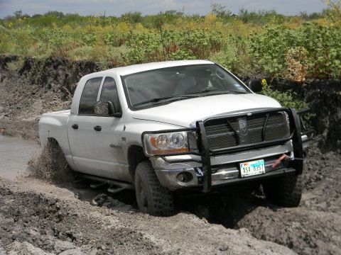 1989 Ford Bronco - This white Dodge Ram pickup truck is navigating a muddy off-road trail, kicking up water and dirt.