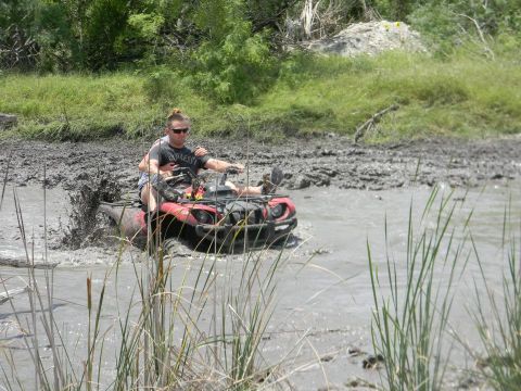 1989 Ford Bronco - Two individuals navigate a red ATV through a muddy, watery terrain, kicking up spray.