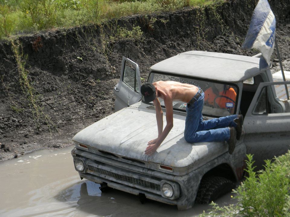 Gray pickup truck stuck in mud with a shirtless man on its hood