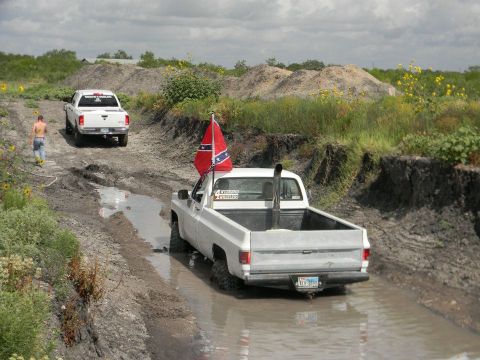 1989 Ford Bronco - Two pickup trucks navigate a muddy off-road course under a cloudy sky.