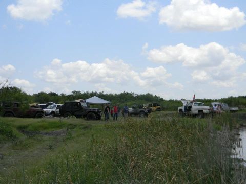 1989 Ford Bronco - A group of off-road vehicles are gathered in a field with a body of water nearby.
