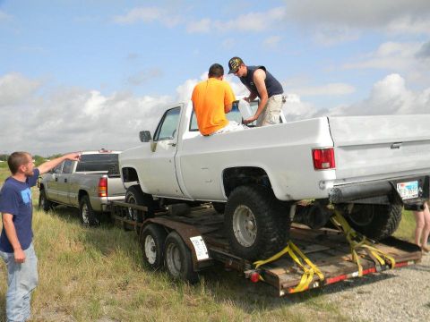 1989 Ford Bronco - A white pickup truck is being transported on a trailer, with two people in the bed and another person pointing from the side.