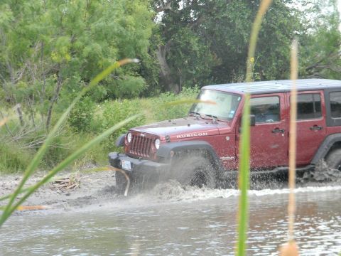 1989 Ford Bronco - A red Jeep Wrangler Rubicon navigates a muddy, watery terrain, kicking up spray as it drives through the obstacle.