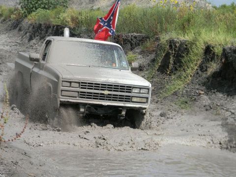 1989 Ford Bronco - Pickup truck navigates a muddy terrain, kicking up water and mud. The grille indicates a 1985 model year.