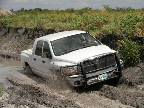 1989 Ford Bronco - A white pickup truck navigates a muddy off-road course, kicking up water and dirt.