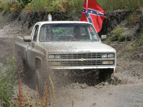 1989 Ford Bronco - A white pickup truck churns through mud, kicking up spray as a confederate flag waves in the background.