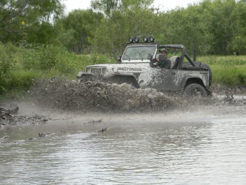 1989 Ford Bronco - A white Jeep Wrangler navigates a muddy off-road course, kicking up a large spray.