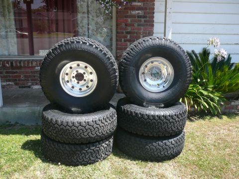 1988 Ford Bronco - Four off-road tires and rims are stacked on grass in front of a house.