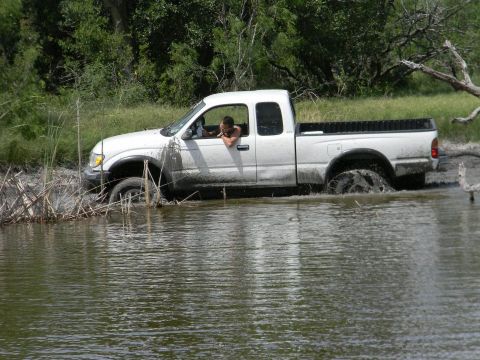 1989 Ford Bronco - A white pickup truck is stuck in murky water, with vegetation on the banks.