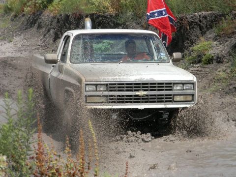1989 Ford Bronco - This lifted pickup truck navigates a muddy terrain, a prominent Chevrolet grille and a Confederate flag waving in the background.