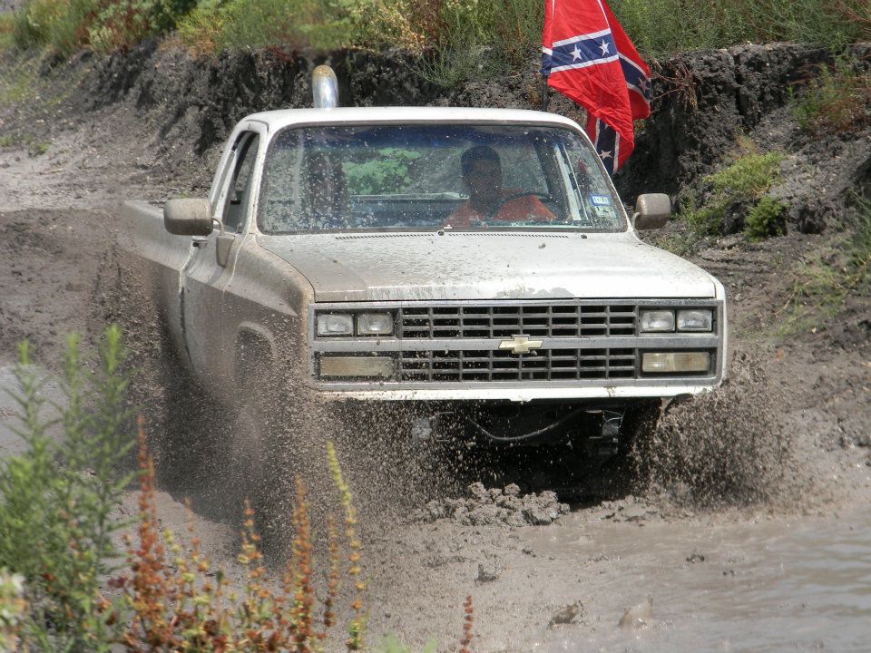White pickup truck with a lifted suspension driving through mud, displaying a Chevrolet emblem and a Confederate flag