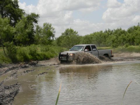 1989 Ford Bronco - A silver pickup truck navigates a muddy, watery terrain, kicking up a large spray.