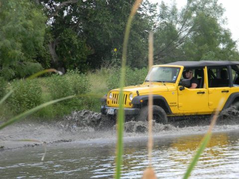 1989 Ford Bronco - A yellow Jeep Wrangler navigates a water obstacle in a natural setting.