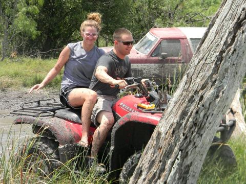 1989 Ford Bronco - Two people ride a red ATV through mud, with a red pickup truck partially visible behind them.