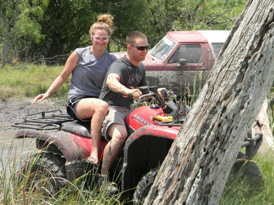 Red ATV with two people covered in mud, with a red pickup truck in the background