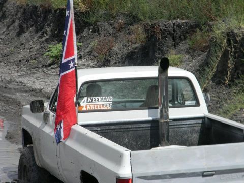 1989 Ford Bronco - A white pickup truck with a tall exhaust pipe and a confederate flag navigates a muddy terrain, with a '4 Weekend Wheelers' sticker on the…