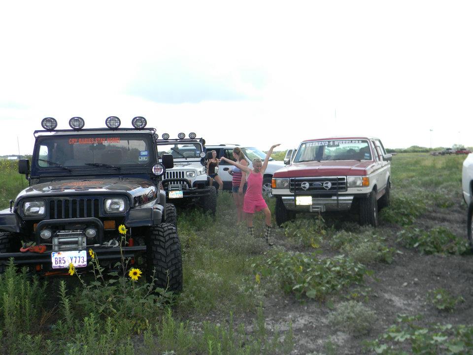 Several Jeeps and a red and white Ford truck parked in a grassy area with people standing around