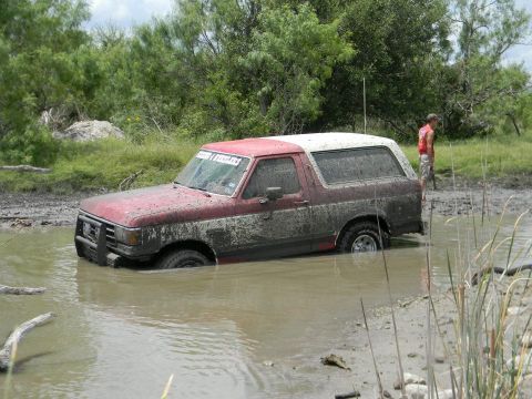 1989 Ford Bronco - A red and white Ford Bronco is partially submerged in muddy water, with a person standing in the background.