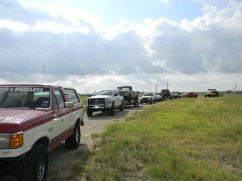 1989 Ford Bronco - A line of trucks and SUVs, including a red and white 1989 Ford Bronco, on a dirt road under a cloudy sky.
