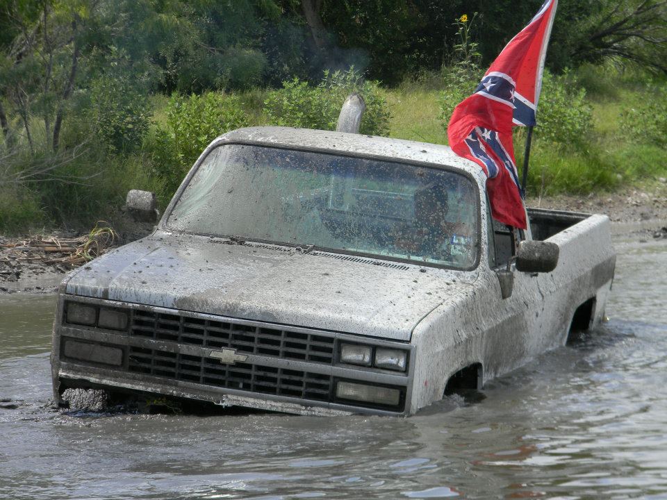 Mud-covered pickup truck stuck in water, Confederate flag flying from the bed. Grille indicates 1985 model