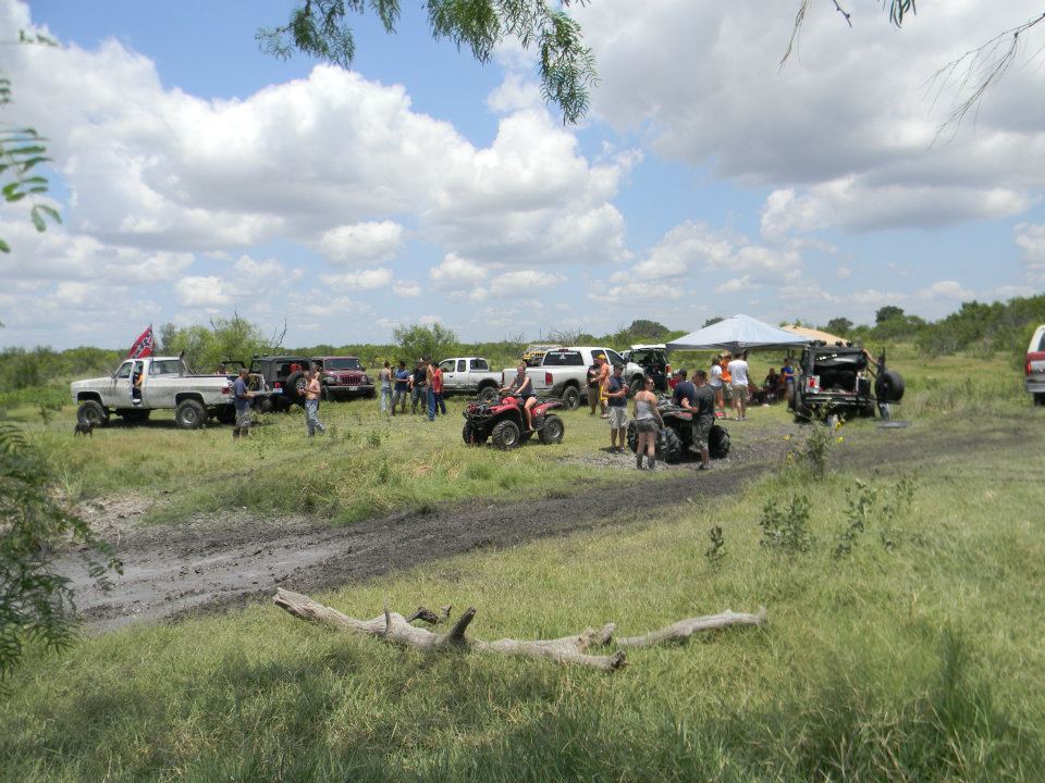 Group of people and several trucks, ATVs, and a buggy gathered in a muddy, grassy field under a cloudy sky
